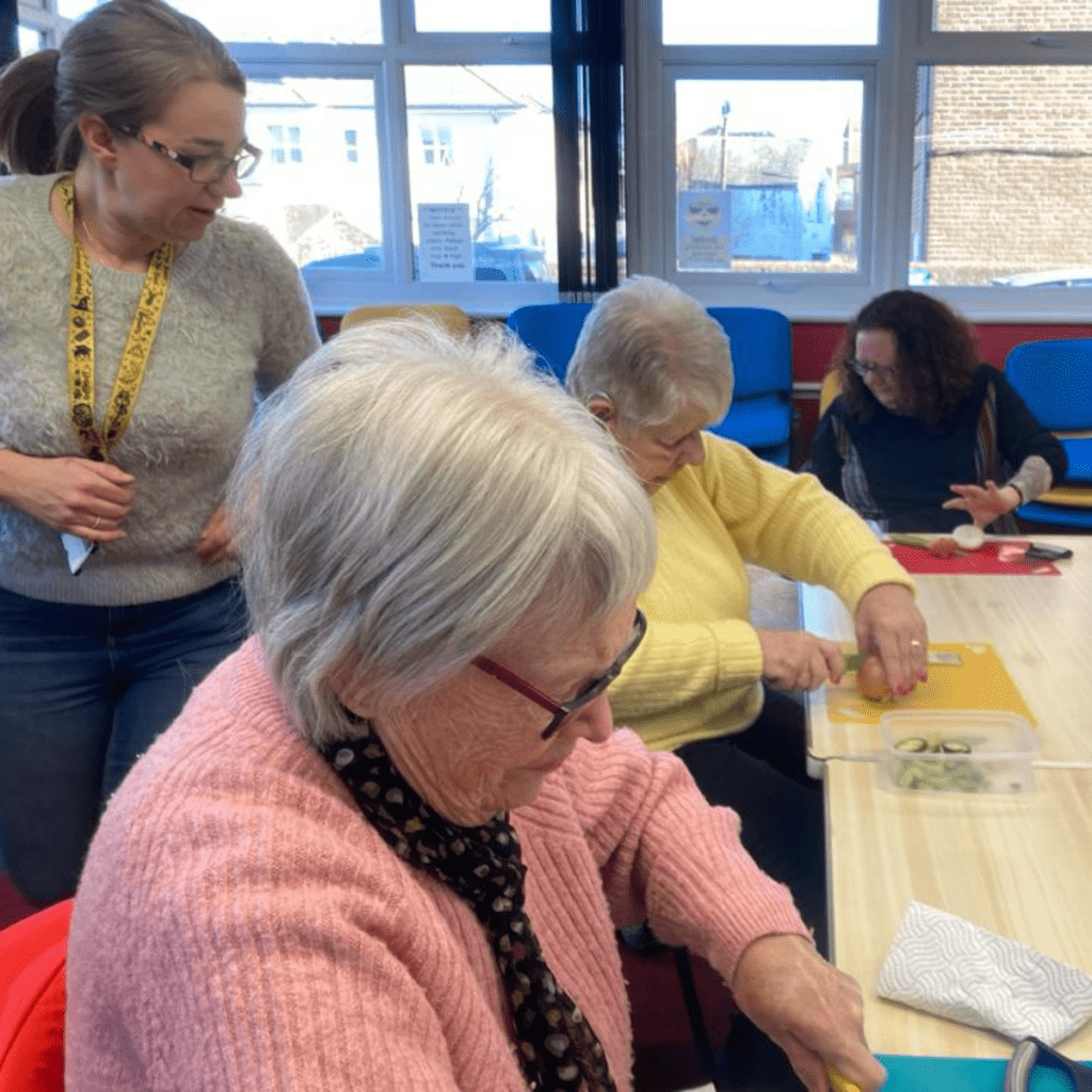 Two women cutting in the cooking lesson with a teacher standing behind them.